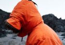 Person i oransje regnjakke med hette i regnvær ved en steinete kyst