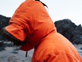 Person i oransje regnjakke med hette i regnvær ved en steinete kyst