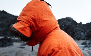Person i oransje regnjakke med hette i regnvær ved en steinete kyst