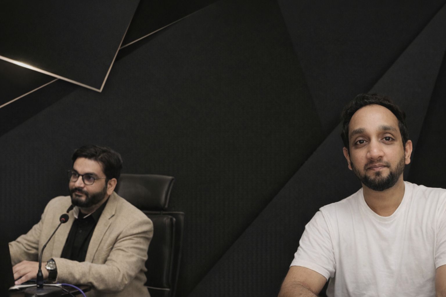 To menn foran en mørk bakgrunn, den ene med mikrofon, den andre ser rett frem.